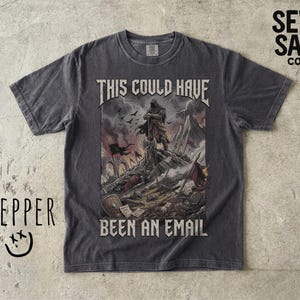 Pode incluir: Uma t-shirt cinzento-carvão de manga curta com um gráfico de um guerreiro num campo de batalha e o texto "THIS COULD HAVE BEEN AN EMAIL" impresso numa fonte desgastada. O design inclui um céu escuro e tempestuoso com destroços.