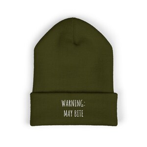 May include: Olive green beanie with the text "WARNING: MAY BITE" in white. The beanie is a solid color and has a folded brim. This hat is a fun accessory.