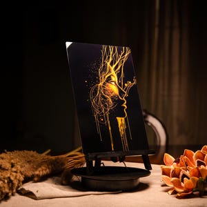 Può includere: Una stampa artistica nera su un piccolo cavalletto, raffigurante un profilo dorato del volto di una donna con linee fluenti. L'opera d'arte è illuminata, creando un contrasto sorprendente con lo sfondo scuro. Fiori decorativi e piante secche in primo piano.