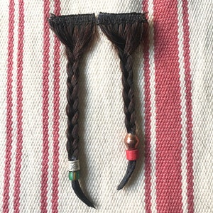 May include: Two dark brown braided hair extensions with black tips. One braid has a silver bead with green and white accents, the other has a copper bead with a red accent. The extensions are on a white and red striped fabric.