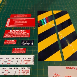 May include: A set of warning labels with black and yellow stripes, red and white text, and a diagram of an electrical circuit. The labels warn of high voltage and electric shock hazards. The text reads "Danger High Voltage 1KV Electric Shock Hazard Remove Power Before Repair or Adjustment".