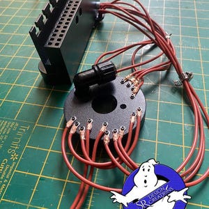 May include: A close-up of a black electrical component with red wires connected to a circular base. The base has multiple small copper contacts with red wires attached. The component is on a green cutting mat with a white grid pattern. The text "SR DESIGN CREATIONS" is visible in a blue circle with a white ghost.