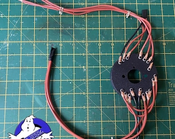 Puede incluir: Primer plano de un arnés de cableado rojo y negro con un conector circular negro. El conector tiene múltiples terminales de cobre. El arnés de cableado está dispuesto sobre una estera de corte verde con marcas en pulgadas. El texto "AF-ASSEMBLED #2" es visible en la parte superior de la imagen.