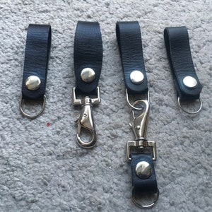 Pode incluir: Quatro chaveiros de couro preto com fechos e argolas de metal prateado.