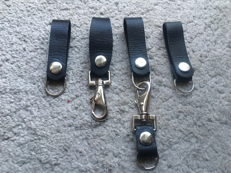 Pode incluir: Quatro chaveiros de couro preto com fechos e argolas de metal prateado.