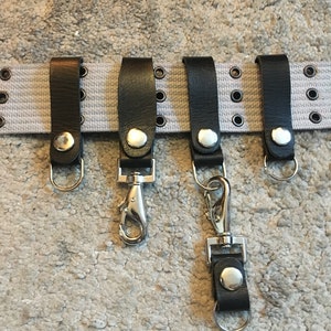 Pode incluir: Cinco chaveiros de couro preto com ganchos e argolas de metal prateado. Os chaveiros est&atilde;o presos a um cinto de lona branco com ilh&oacute;s de metal prateado.