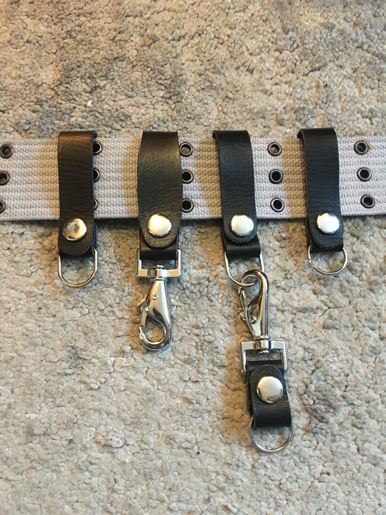 Pode incluir: Cinco chaveiros de couro preto com ganchos e argolas de metal prateado. Os chaveiros est&atilde;o presos a um cinto de lona branco com ilh&oacute;s de metal prateado.