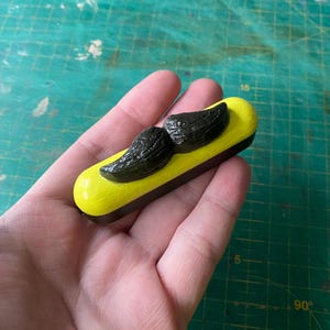 Puede incluir: Un pequeño contenedor amarillo y negro con un diseño de bigote negro. El contenedor mide aproximadamente 7,5 cm de largo y 2,5 cm de ancho.