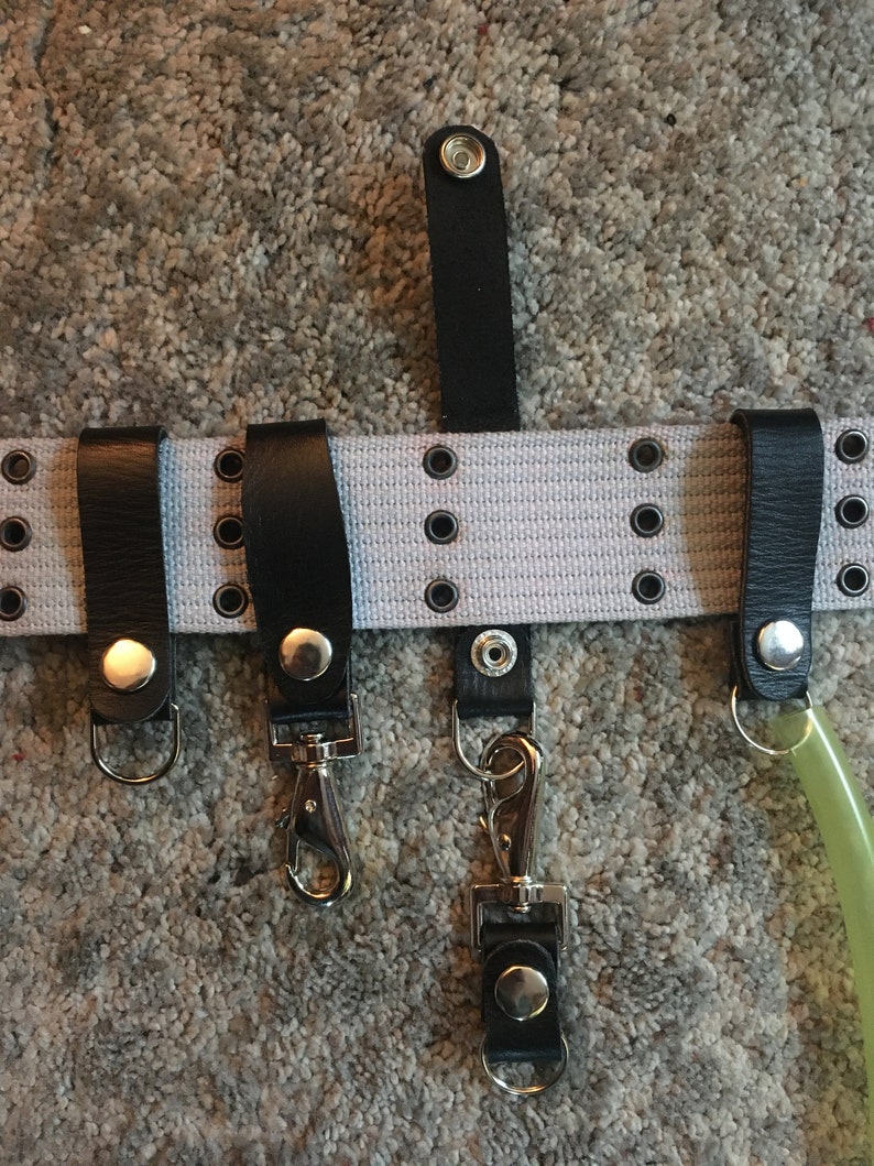 Pode incluir: Um cinto de lona branco com la&ccedil;os de couro preto e fechos de press&atilde;o. O cinto tem v&aacute;rias filas de ilh&oacute;s prateados. H&aacute; tr&ecirc;s chaveiros prateados presos aos la&ccedil;os. Um chaveiro tem um gancho prateado, outro tem um mosquet&atilde;o prateado e o terceiro tem um anel prateado.