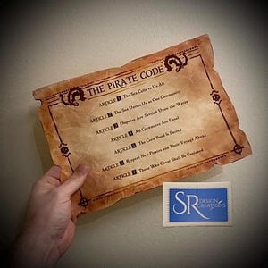 May include: Aged parchment-style print of "The Pirate Code" with decorative borders and text. The text includes articles about community, equality, and punishment. A small blue and white logo is in the lower right corner.