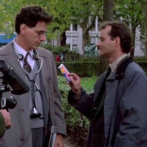 May include: Two men standing in a park, one wearing a grey coat and a stethoscope, the other wearing a blue coat and holding a candy bar.