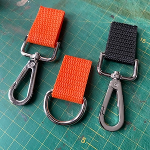 Puede incluir: Dos clips metálicos con correas de tela naranja y negra. Los clips tienen un acabado plateado y una forma de D. Las correas están hechas de tela tejida.