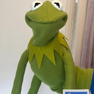 May include: A green felt frog puppet with large white eyes, black pupils, and a pointed collar. The puppet features a wide, smiling mouth and long, slender arms. A small white rectangular tag with blue text reading SR DESIGN CREATIONS is attached.