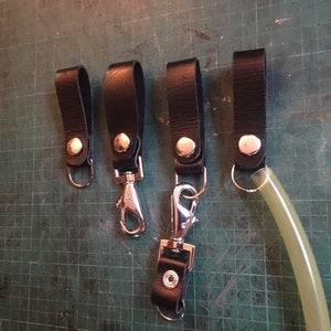 Pode incluir: Cinco chaveiros de couro preto com fechos e argolas de metal prateado. Os chaveiros t&ecirc;m um &uacute;nico bot&atilde;o prateado cada um.