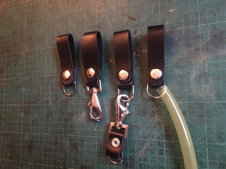 Pode incluir: Cinco chaveiros de couro preto com fechos e argolas de metal prateado. Os chaveiros t&ecirc;m um &uacute;nico bot&atilde;o prateado cada um.