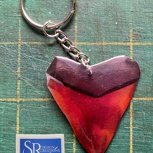 May include: A red and black shark tooth keyring with a silver key ring. The keyring is made of resin and has a glossy finish. The tooth is shaped like a heart and has a small hole at the top for the key ring. The keyring is on a green cutting mat with a white label that says "SR Design Creations".