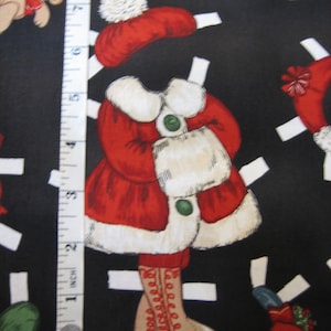 May include: A black fabric featuring Christmas-themed paper dolls. The dolls are dressed in red coats with white fur trim, matching hats, and boots. A measuring tape is placed vertically along the left side of the fabric, showing inches.