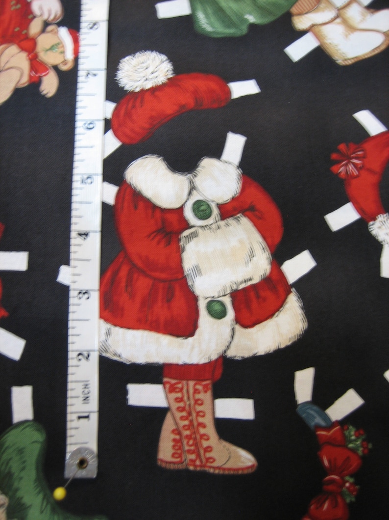 May include: A black fabric featuring Christmas-themed paper dolls. The dolls are dressed in red coats with white fur trim, matching hats, and boots. A measuring tape is placed vertically along the left side of the fabric, showing inches.