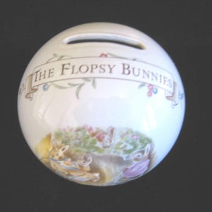 May include: White ceramic piggy bank shaped like a sphere with a slot on top. The bank features a colorful illustration of the characters from the book "The Flopsy Bunnies" by Beatrix Potter.