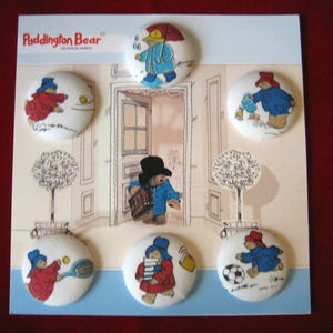 Może przedstawiać: Zestaw przypinek z misiem Paddington na karcie ekspozycyjnej. Przypinki przedstawiają misia Paddington w różnych sytuacjach, takich jak gra w tenisa, noszenie książek i gra w piłkę nożną. Tło zawiera rysunek drzwi.