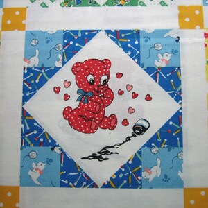 Op de afbeelding: Een kleurrijk quiltvierkant met een centrale ruit met een rode, gestippelde beer die zijn handen tegen zijn borst houdt, omringd door harten. De beer staat op een witte achtergrond, met een gemorste inktfles. De quilt heeft een blauw, wit, geel en groen patroon.