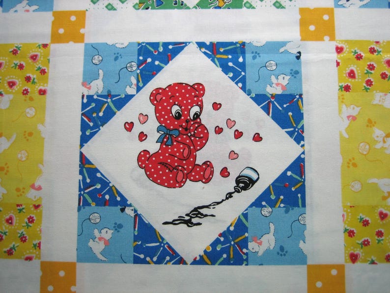 Op de afbeelding: Een kleurrijk quiltvierkant met een centrale ruit met een rode, gestippelde beer die zijn handen tegen zijn borst houdt, omringd door harten. De beer staat op een witte achtergrond, met een gemorste inktfles. De quilt heeft een blauw, wit, geel en groen patroon.