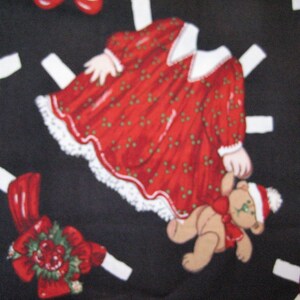 May include: A fabric pattern featuring Christmas-themed paper dolls on a black background. The dolls include a red dress with white lace trim, a teddy bear, and other festive elements. The design is whimsical and holiday-themed.