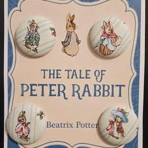 Puede incluir: Cuatro botones redondos con ilustraciones de los personajes de Peter Rabbit de la historia de Beatrix Potter, "El cuento de Peter Rabbit". Los botones están colocados sobre un fondo crema con detalles azules y texto.