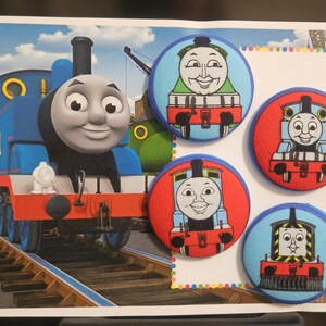 Könnte beinhalten: Ein Bild mit dem blauen Thomas, der Tank Engine, und vier runden Knöpfen. Die Knöpfe zeigen verschiedene Zugcharaktere auf roten oder blauen Hintergründen. Der Hintergrund zeigt eine Bahngleis und eine grüne Lokomotive.
