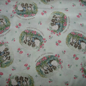 May include: Light blue fabric with a repeating pattern of illustrations of three kittens surrounded by a floral wreath. The text "THREE LITTLE KITTENS" is printed around each illustration. Small red flowers and green leaves are scattered across the fabric.