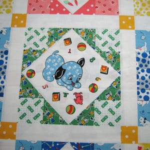 Op de afbeelding: Een patchworkquilt met een centraal vierkant met een blauwe puppy-illustratie omringd door kleurrijke vormen en cijfers. De quilt heeft een verscheidenheid aan patronen in blauw, groen, geel en rood, wat een speelse en vintage esthetiek cre&euml;ert.