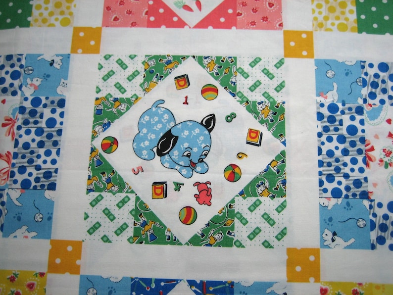 Op de afbeelding: Een patchworkquilt met een centraal vierkant met een blauwe puppy-illustratie omringd door kleurrijke vormen en cijfers. De quilt heeft een verscheidenheid aan patronen in blauw, groen, geel en rood, wat een speelse en vintage esthetiek cre&euml;ert.