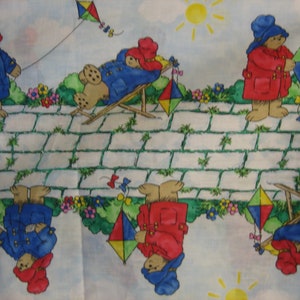 以下が含まれることがあります： 赤と青のコートを着たクマが繰り返し描かれたカラフルな生地。クマたちは凧を持ち、ビーチチェアでリラックスしています。背景は白いレンガの道で、緑の草と花が咲いています。