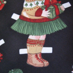 May include: A paper doll illustration of a child in a Christmas outfit. The child wears a tan sweater with green Christmas trees, a green skirt, and red-trimmed socks and boots. The child holds a green gift box with a red bow.