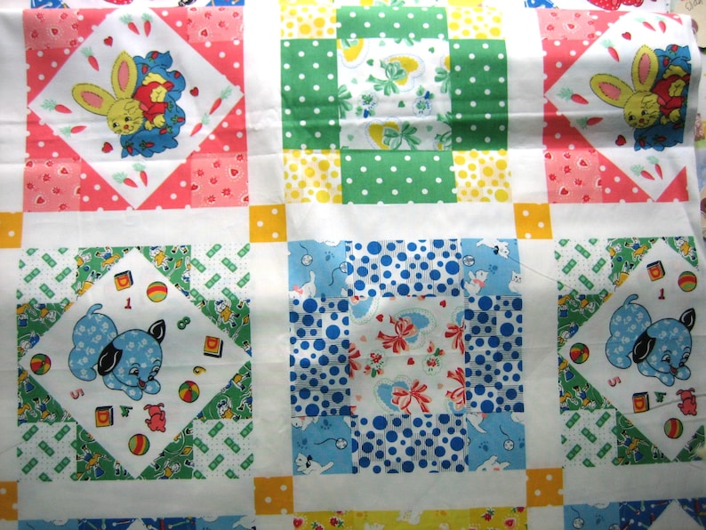Op de afbeelding: Een patchworkquilt met vierkanten met verschillende patronen en afbeeldingen. De quilt toont een herhalend patroon van vierkanten met een gele konijn, een blauwe puppy en stippen in groen en blauw. De quilt is gemaakt van zachte stof.