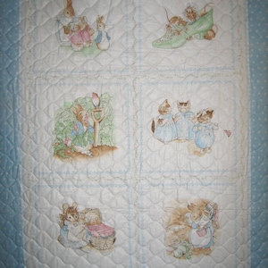 Könnte beinhalten: Eine weiße gesteppte Babydecke mit blauem Rand und weißen Punkten. Die Decke zeigt sechs verschiedene Szenen aus den Peter Rabbit-Geschichten, darunter Peter Rabbit, Benjamin Bunny und die drei kleinen Kaninchen.