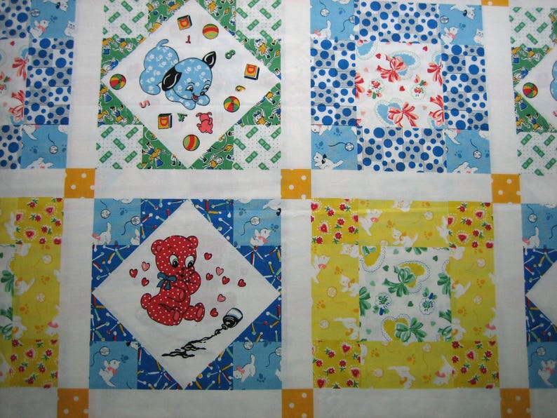 Op de afbeelding: Een patchworkquilt met een herhalend patroon van vierkanten. Elke vierkant heeft een ander ontwerp, waaronder een blauwe puppy, een rode teddybeer en bloemen- en stippenpatronen. De quilt is voornamelijk wit, blauw, geel en groen.