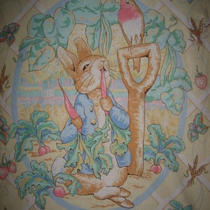 May include: A fabric featuring a circular illustration of Peter Rabbit eating a carrot, surrounded by garden elements. A robin sits on a shovel. The design includes strawberries, carrots, and radishes, with the text "Peter Rabbit" at the bottom.