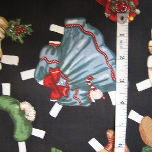 May include: A black fabric featuring a repeating pattern of Christmas-themed paper dolls. The dolls include a blue dress with a red bow, a green hat, and a dog wearing a plaid scarf. A measuring tape shows a length of approximately 3 inches.