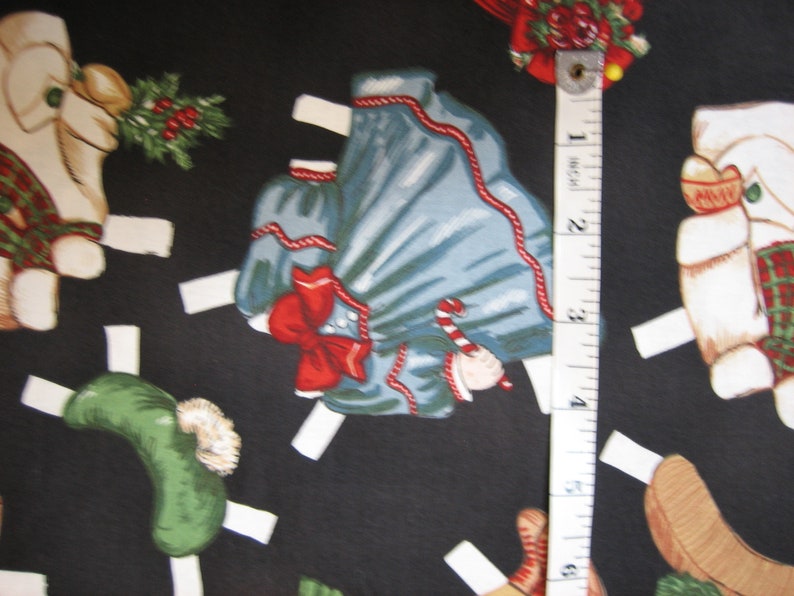 May include: A black fabric featuring a repeating pattern of Christmas-themed paper dolls. The dolls include a blue dress with a red bow, a green hat, and a dog wearing a plaid scarf. A measuring tape shows a length of approximately 3 inches.