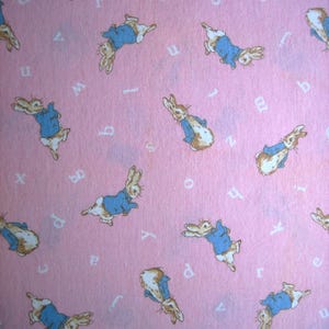 May include: A pink fabric featuring a repeating pattern of Peter Rabbit characters in blue jackets and white undercoats. White letters are scattered across the background.