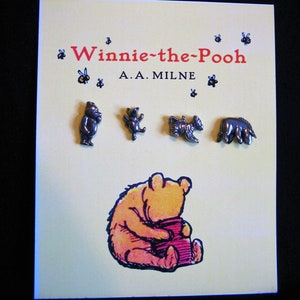 May include: A book cover with the title "Winnie-the-Pooh" and author A.A. Milne. The cover is light yellow with illustrations of bees and Winnie the Pooh characters. Four small metal figurines of the characters are placed above the illustration.