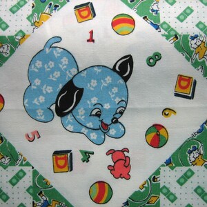 Op de afbeelding: Een vintage quiltvierkant met een lichtblauwe puppy met een bloemenpatroon, zwarte oren en een rode tong. Het ontwerp bevat kleurrijke ballen, blokken, cijfers en een kleine rode hond op een witte achtergrond, omlijst door groen-witte patronen.