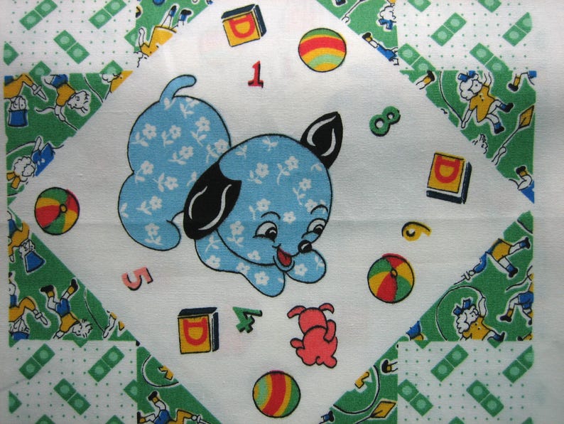 Op de afbeelding: Een vintage quiltvierkant met een lichtblauwe puppy met een bloemenpatroon, zwarte oren en een rode tong. Het ontwerp bevat kleurrijke ballen, blokken, cijfers en een kleine rode hond op een witte achtergrond, omlijst door groen-witte patronen.