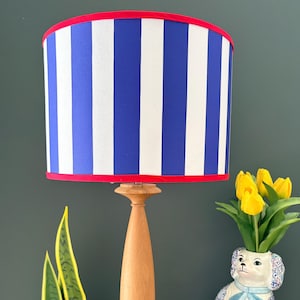 May include: A table lamp with a light wood base and a cylindrical lampshade featuring vertical blue and white stripes with red trim. The lamp is positioned next to a ceramic dog-shaped vase holding yellow tulips and a stack of books.