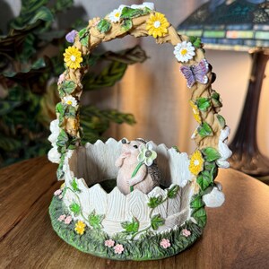 May include: A decorative garden basket with a hedgehog figurine holding a flower. The basket is adorned with yellow and white flowers, green leaves, and a purple butterfly. The base is green with pink flowers.