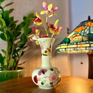 May include: A small, cream-colored vase with a bulbous base and a narrow neck, adorned with pink and red floral designs. The vase holds several stems of orchids with yellow and pink petals. The vase is on a wooden surface.