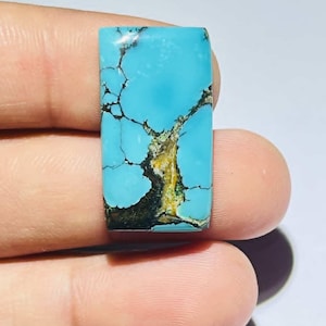 Puede incluir: Gema rectangular de turquesa con una llamativa matriz de vetas marrón oscuro y doradas. La piedra pulida muestra un tono azul vibrante, ideal para la fabricación de joyas o coleccionar. La piedra se sostiene entre los dedos.