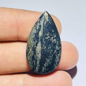 Pode incluir: Uma pedra preciosa em forma de gota com uma superfície polida. A pedra é principalmente preta com raios dourados e brancos. A pedra preciosa é segurada entre os dedos.