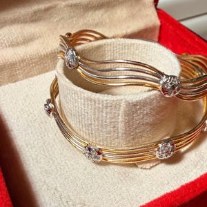 Könnte beinhalten: Zwei Armreifen aus goldfarbenem Metall mit einem mehrsträngigen Design und wellenförmigen Linien. Jeder Armreif ist mit kreisförmigen, silberfarbenen Fassungen verziert, die kleine, klare, funkelnde Steine enthalten, die gleichmäßig verteilt sind.